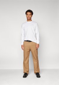 T-shirt branca de mangas longas com uma marcação subtil, combinada com calças de fato castanho claro e sapatos pretos. O modelo está posicionado contra um fundo neutro.