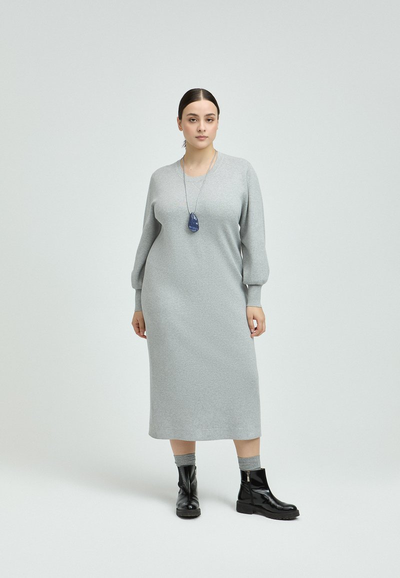 Graues, langärmliges Strickkleid in Midi-Länge mit rundem Ausschnitt. Kombiniert mit schwarzen Stiefeletten und einer blauen Anhänger-Halskette.