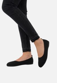 Schwarze Slipper aus strukturiertem Stoff. Rundliche Zehenform, minimalistischer Stil und flache Sohle. Getragen mit eng anliegenden schwarzen Jeans.
