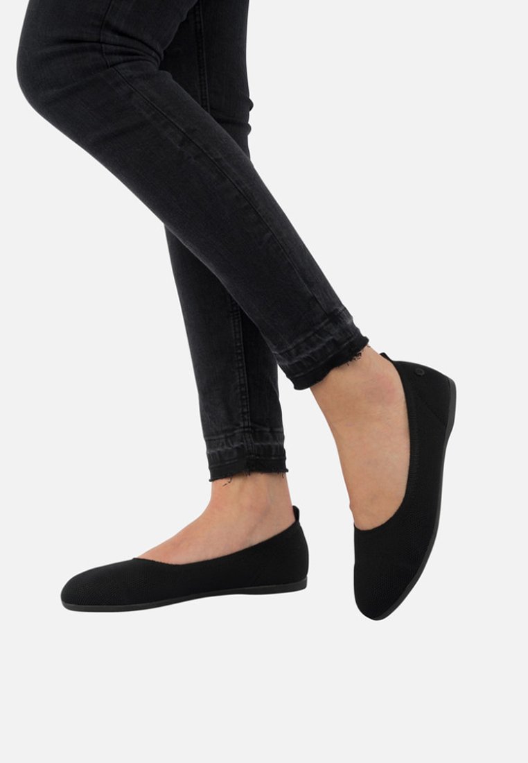 Schwarze Slipper aus strukturiertem Stoff. Rundliche Zehenform, minimalistischer Stil und flache Sohle. Getragen mit eng anliegenden schwarzen Jeans.
