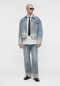 Homme portant une veste en jean délavée et un jean, une chemise blanche, une cravate noire, des chaussures noires et des lunettes de soleil foncées, debout devant un fond uni.