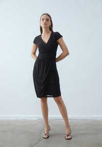Black wrap dress featuring a v-neck, short sleeves, and a textured lace panel on one side, with a flowing silhouette and knee-length hem.