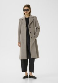 Cappotto di lana beige con colletto a punta, tre bottoni e tasche laterali, abbinato a una maglietta nera e pantaloni a gamba larga. Scarpe nere e occhiali da sole.