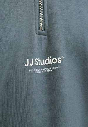 Modrá látka s vertikálnym zipsom nad bielym textom, ktorý znie „JJ Studios, brought to you by the J&J Crew, designed in Denmark.“