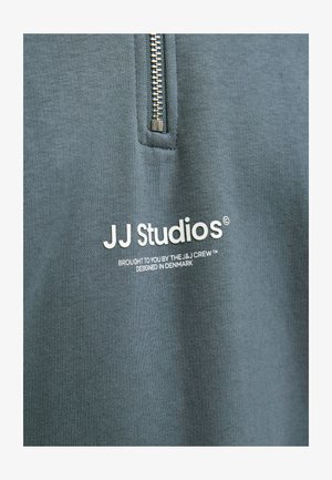 Blauwe stof met een verticale rits boven witte tekst waarop staat: "JJ Studios, gebracht door de J&J Crew, ontworpen in Denemarken."