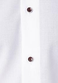 Gros plan d’un tissu blanc avec deux boutons marron foncé cousus sur la patte verticale d’une chemise.