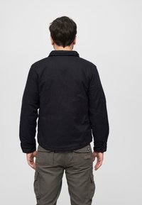 Svart, vadderad jacka med krage, knappmanschetter och rak nederkant. Buren över olivgröna cargobyxor, sedd bakifrån.