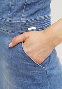 Main placée dans la poche avant d'un jean en denim bleu, montrant le tissu texturé et une petite étiquette métallique de la marque au-dessus de la poche.