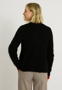 Sweater noir en tricot côtelé avec une coupe décontractée et des manches longues. Présente un accent rouge contrastant aux poignets. Texture douce, épaisseur moyenne.