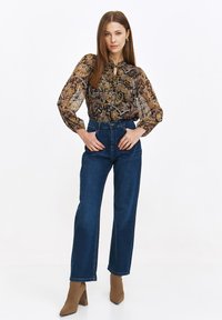 Bluzka w brązowo-złoty wzór z wiązaniem przy szyi, zestawiona z ciemnoniebieskimi dżinsami z wysokim stanem i beżowymi botkami na słupku.