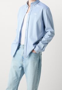 Hellblaues Langarmhemd mit Knopfleiste, versehen mit einem kleinen weißen Logo auf der Brust und einem lockeren Schnitt. Trägt man über einem weißen Tanktop.