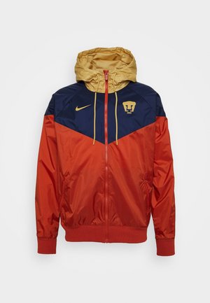Rode, marineblauwe en gouden windjack met capuchon en rits, met Nike-logo en gouden Puma UNAM teampatch op de borst.