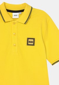 Žluté polo tričko s černým lemovaným límcem, se třemi knoflíky a čtvercovou nášivkou "BOSS" na levé části hrudi.