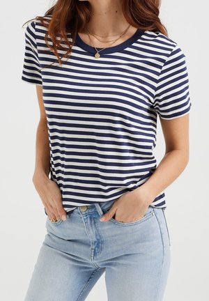 Vrouw draagt een marineblauw-wit gestreept T-shirt met korte mouwen, lichtblauwe spijkerbroek, gouden ketting en ringen, met handen in de zakken en zichtbaar rood haar.
