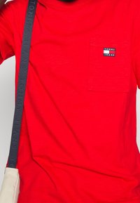 Rød kortærmet t-shirt med en venstre brystlomme, der har en sort lap med teksten "TOMMY JEANS." Mønstrede stoffer og en rund halsudskæring.