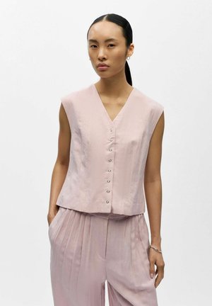Donna con capelli neri lisci raccolti in una coda bassa, indossa un top rosa pallido senza maniche con bottoni e pantaloni plissettati abbinati, mani nelle tasche.
