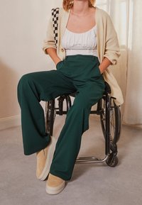 Green wide-leg trousers with a high waist, paired with a white top and beige slip-on shoes. A beige knitted cardigan adds texture.