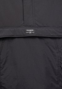 Sort nylonjakke med lynlås, sidelommer og et mærke med "PULL&BEAR" og "Designet i Europa." Glat tekstur.