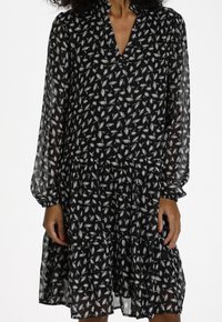 Svart klänning med ett blommigt vitt mönster, långa genomskinliga ärmar med elastiska manschetter, och en flikig kjoldesign.