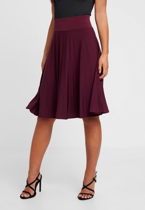 Femme portant une jupe plissée bordeaux mi-longue associée à des sandales à talons hauts noires à brides sur un fond uni.