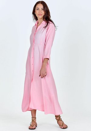 Donna che indossa un vestito rosa a maniche lunghe con bottoni e sandali marroni con cinturini, in piedi su uno sfondo bianco.