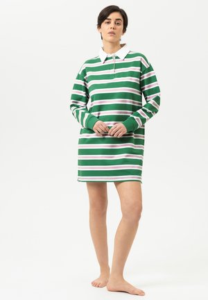 Frau steht barfuß und trägt ein langärmliges Kleid mit horizontalen grünen, weißen und pinken Streifen und einem weißen Kragen vor einem schlichten Hintergrund.