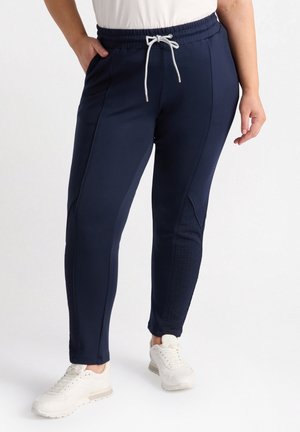 Persoon draagt marineblauwe joggers met elastische tailleband en wit koord, gecombineerd met witte sneakers, staand tegen een effen achtergrond.