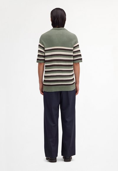 Persona in piedi di spalle che indossa una maglia a maniche corte a righe verdi e marroni in maglia e pantaloni larghi scuri con scarpe nere.