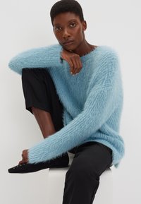 Helles bläuliches, flauschiges Sweatshirt mit legerem Schnitt, strukturiertem Strick und langen Ärmeln, kombiniert mit schwarzen, kürzeren Hosen und schwarzen Schuhen.