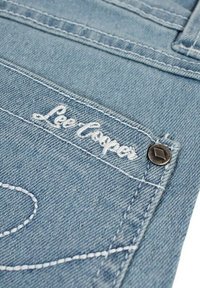 Tissu en denim bleu clair avec des surpiqûres blanches, arborant un logo Lee Cooper en écriture cursive et un détail de bouton en métal noir.