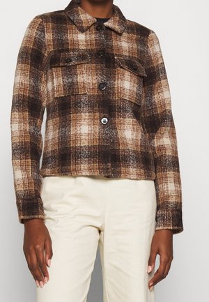 Brown and beige plaid button-up jacket with flap chest pockets worn with off-white pants on a standing person.
