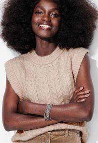 Pull sans manches en tricot beige avec un motif en torsades, doté d'un col rond, associé à un bracelet en argent empilé au poignet.