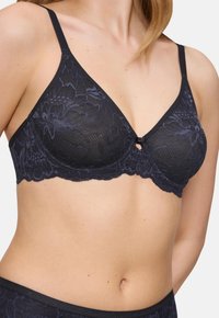 Žena nosi crni čipkasti grudnjak s cvjetnim uzorcima, tankim trakama i malim mašnom na prednjem dijelu.