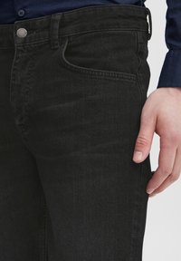 Crne traperice od denima slim kroja, s prednjim džepom, metalnim zatvaranjem na dugme i suptilnim detaljima šivanja na džepovima.