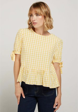 Femme portant un chemisier manches courtes à carreaux jaune et blanc avec manches nouées et ourlet à volants, associé à un jean bleu foncé.