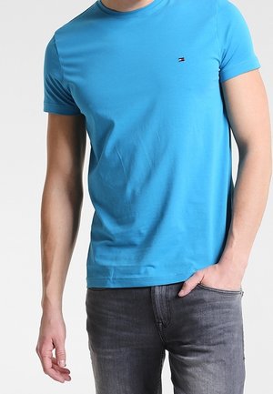 Mann trägt ein enganliegendes, leuchtend blaues kurzärmliges T-Shirt mit einem kleinen Tommy Hilfiger-Logo auf der Brust und dunkelgrauen Jeans.