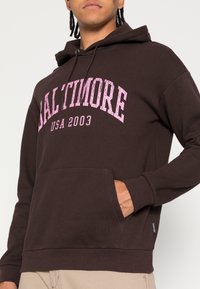 Hnědá mikina s kapucí, přední kapsou a šňůrkou na stažení, s bílým a růžovým opotřebovaným nápisem "BALTIMORE USA 2003" na hrudi.