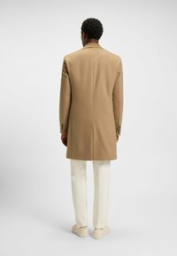 Cappotto di lana beige con design su misura, maniche lunghe e spacco posteriore. Abbinato a pantaloni bianchi e sneaker beige. Sfondo semplice.