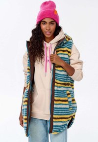 Mitmevärviline fliisvest, millel on sinised, kollased ja pruunid triibud, lukuga kinnitamine, koos kreemja kapuutsiga ja roosa mütsiga. Igapäevane disain.