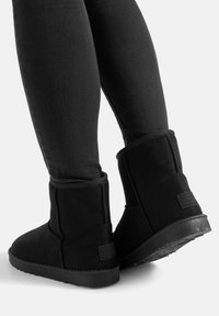 Persona che indossa stivali neri a metà polpaccio con suole spesse e leggins neri, in piedi su sfondo bianco.