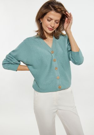 Lichtblauwe geribbelde cardigan met een V-hals, voorzien van houten knopen aan de voorkant. Combinatie met witte broek die een ontspannen pasvorm heeft.