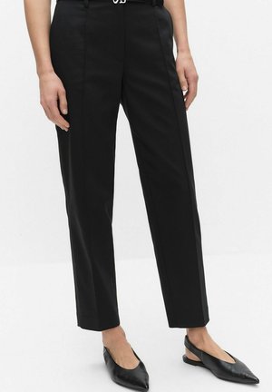 Le bas du corps d'une personne portant un pantalon noir sur mesure avec une ceinture et des chaussures plates noires à bout pointu et bride arrière, debout devant un fond blanc uni.