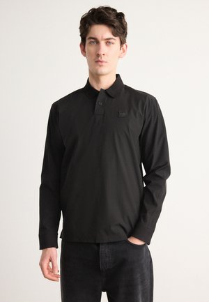 Polo-shirt noir à manches longues avec col à boutons, en tissu lisse. Présente un petit patch logo sur la poitrine gauche. Associé à un jean foncé.