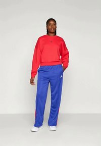 Sarkans sporta krekls ar apaļu kaklu, brīvu piegriezumu un Adidas logo. Zili sporta bikses ar sānu svītrām un elastīgu jostu. Baltas sporta apavi.