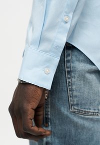 Χέρι με σκούρο δέρμα που φορά αλυσίδα από ασήμι, μανσέτα γαλάζιου πουκάμισου με λευκά κουμπιά και κεντημένο λογότυπο CK, δίπλα στην τσέπη μπλε τζιν.