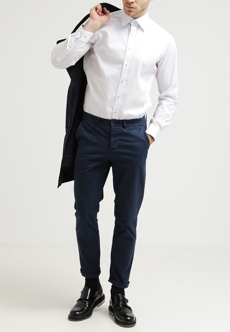 Homme portant une chemise blanche boutonnée rentrée dans un pantalon bleu marine, tenant une veste noire sur l'épaule, debout devant un fond uni.