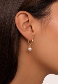 Boucles d'oreilles en or en forme de hoop avec une goutte de perle, présentant une finition brillante et une texture lisse, suspendues à un petit lien en or.