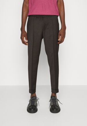 Hombre con pantalones a medida color marrón oscuro con puños y zapatillas negras con cordones blancos con estampado, de pie frente a un fondo blanco.