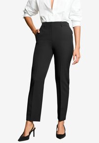 Una persona indossa una camicia bianca elegante e pantaloni neri attillati, in piedi con sicurezza con una mano nella tasca, accentuata da tacchi neri.
