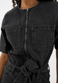 Čierny denimo overal s krátkymi rukávmi, predným zipsom, dvoma náprsnými vreckami a viazaním v páse. Obsahuje vyblednutú textúru a kontrastné šitie.